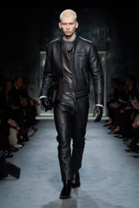 Male model with platinum hair walks a fashion runway in a black leather biker outfit and gloves, carrying a clutch under his arm.