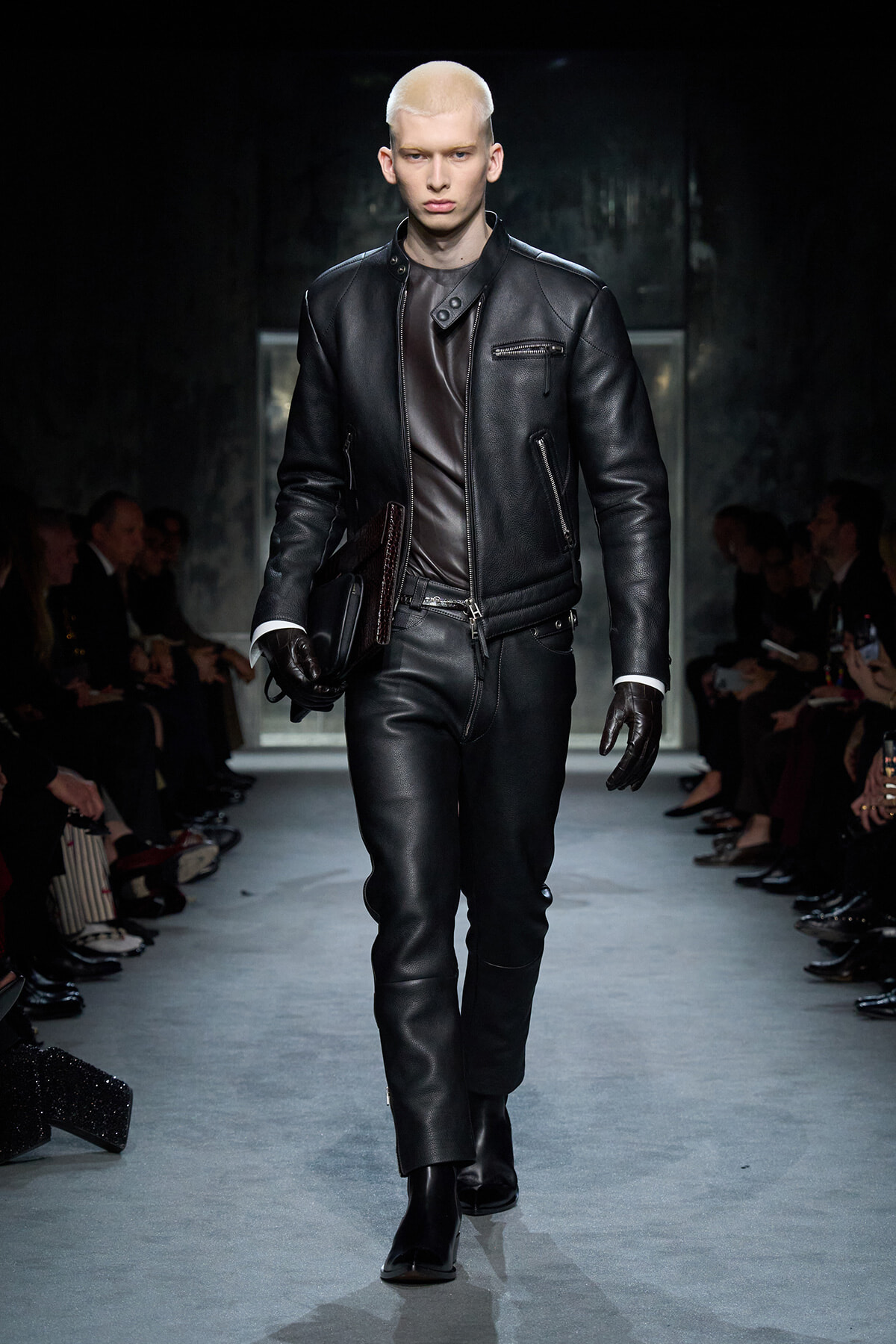 Male model with platinum hair walks a fashion runway in a black leather biker outfit and gloves, carrying a clutch under his arm.