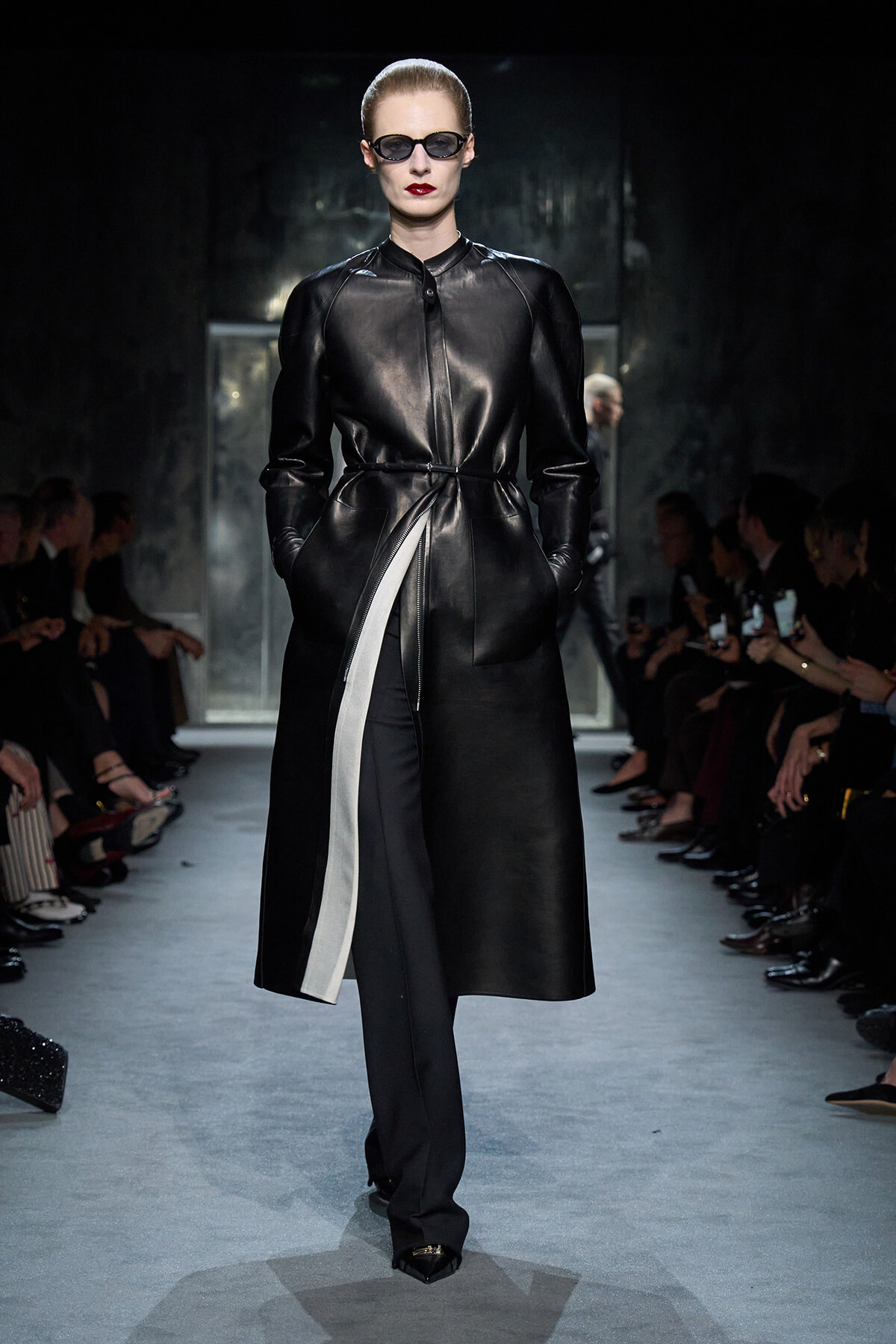 Model on a runway wearing a black leather trench coat with a belt, dark sunglasses, and bold red lipstick.