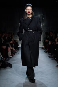 Model walks the runway in a long black double-breasted coat with a high, brown leather collar and belt at the waist.