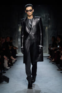 Male model on a fashion runway wearing a long black leather coat, black gloves, and dark sunglasses.