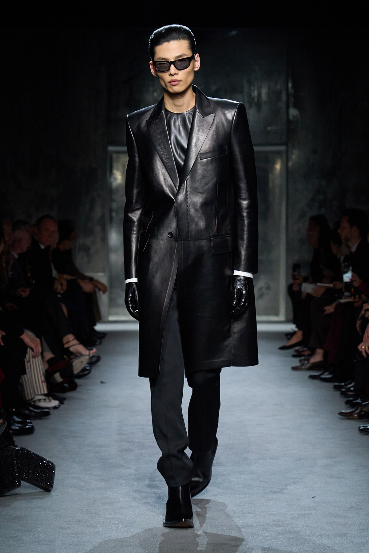 Male model on a fashion runway wearing a long black leather coat, black gloves, and dark sunglasses.