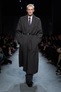 Male model on a runway wearing a long dark overcoat belted at the waist, pink shirt and dotted tie, with audience on both sides.