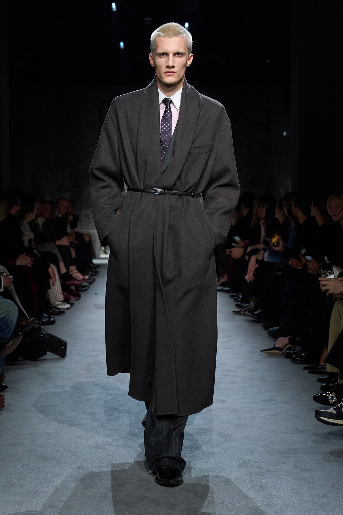 Male model on a runway wearing a long dark overcoat belted at the waist, pink shirt and dotted tie, with audience on both sides.