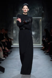 Model in a black, floor-length knit dress with a thin belt and dark leather gloves, standing with arms crossed on a runway.