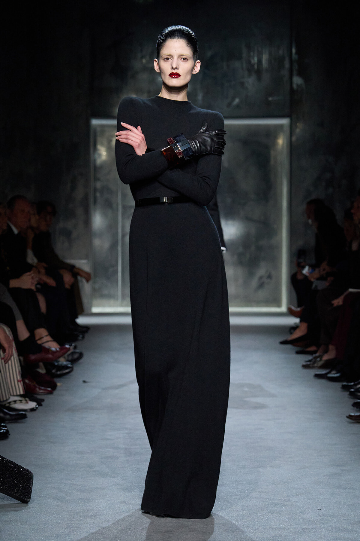 Model in a black, floor-length knit dress with a thin belt and dark leather gloves, standing with arms crossed on a runway.