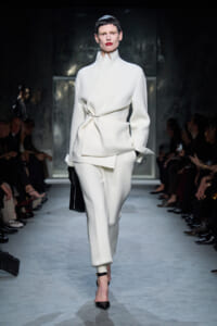 Model on a runway wearing a white sculptural suit with a wrapped waist, matching pants, black gloves, and a black clutch.