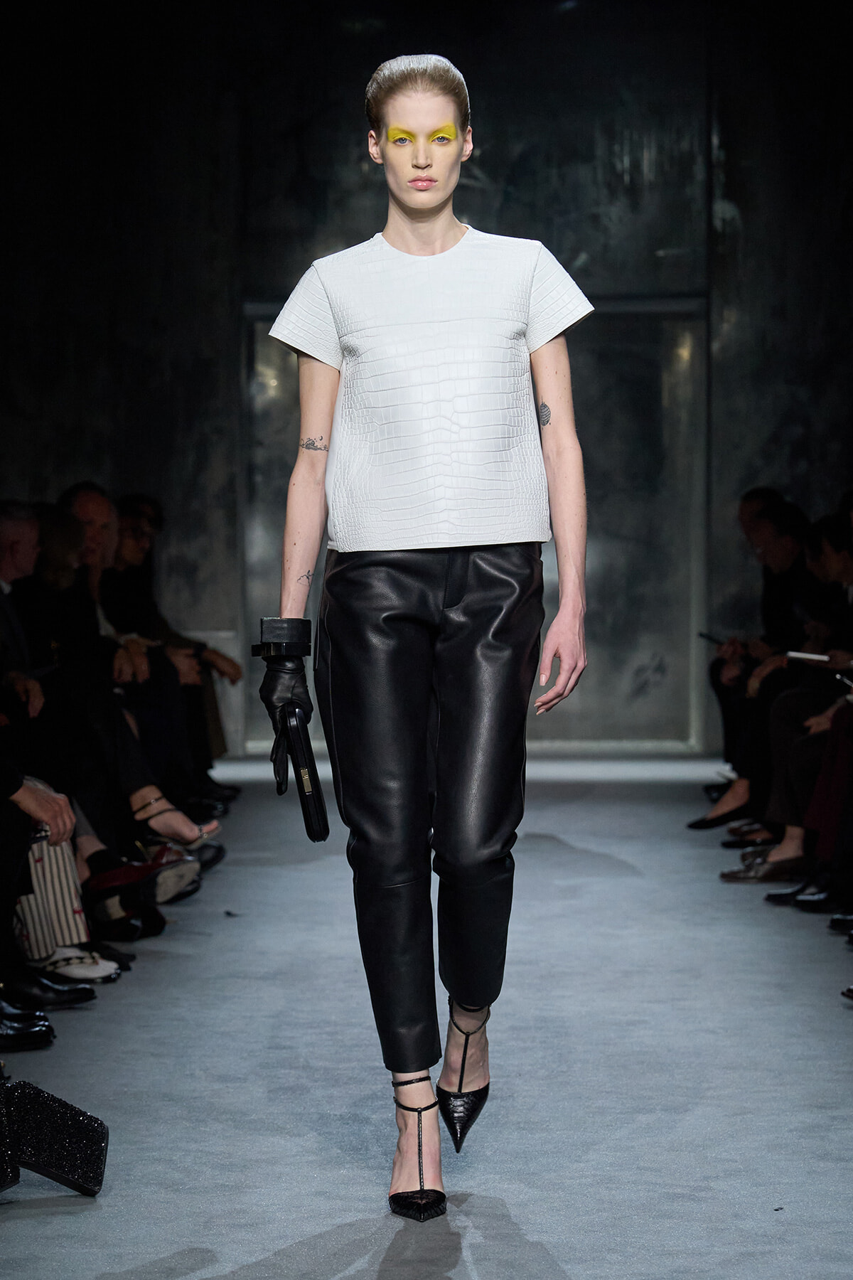 Model on a runway wearing a white textured short-sleeve top and black leather pants with yellow eye makeup and pointed black heels, carrying a black clutch glove on one hand.