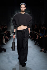 Model on a runway wearing a cropped black sweater and wide-leg black trousers, carrying a glossy black handbag.