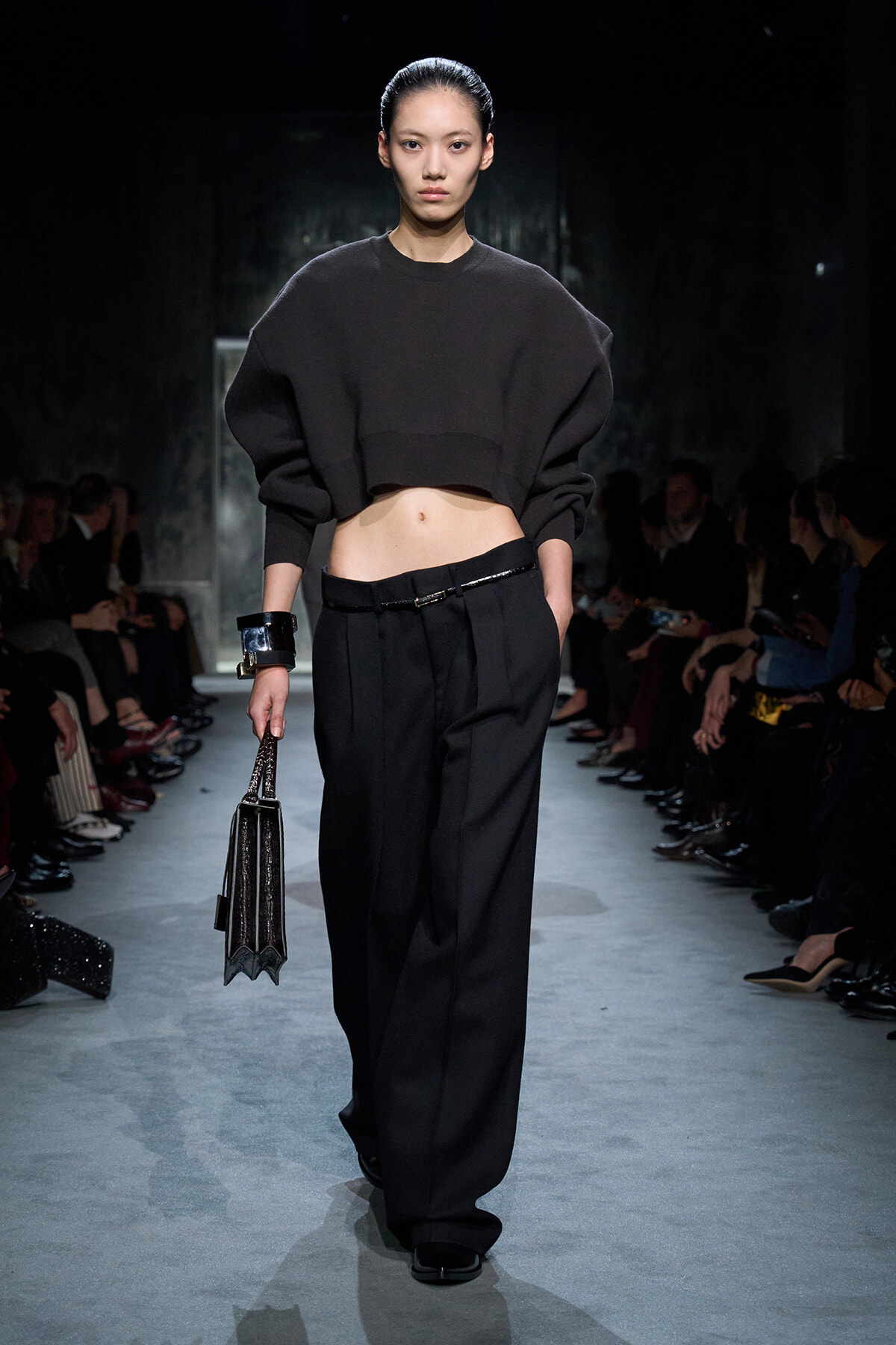 Model on a runway wearing a cropped black sweater and wide-leg black trousers, carrying a glossy black handbag.