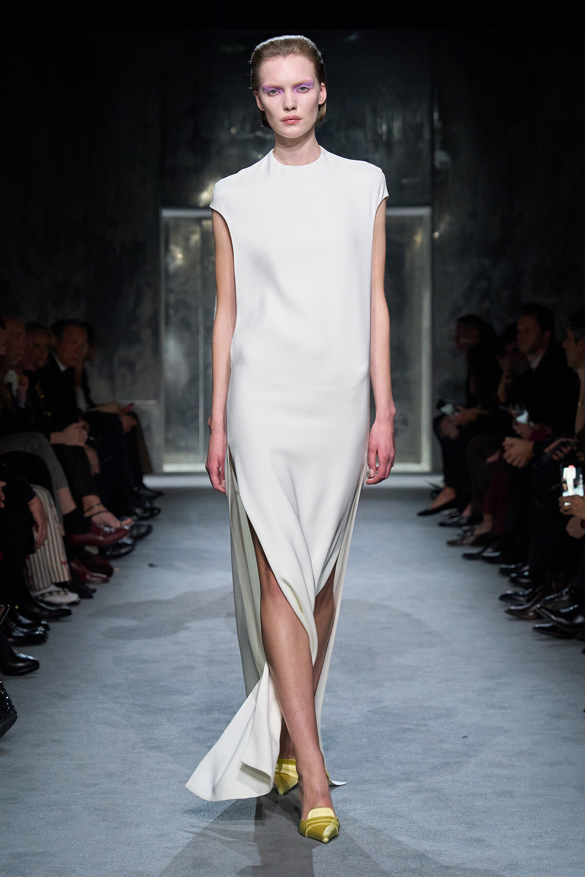 Model walks a fashion runway in a sleeveless white column dress with a high front slit, paired with yellow flats.