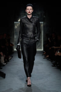 Model wearing a black leather biker jacket and matching pants walks the runway at a fashion show with a clutch in hand.