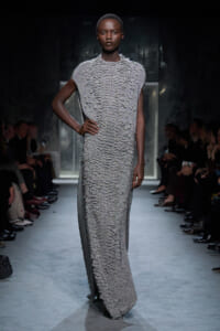 Model on a runway wearing a shimmering, textured silver floor-length gown with short sleeves, audience lining the catwalk on both sides.