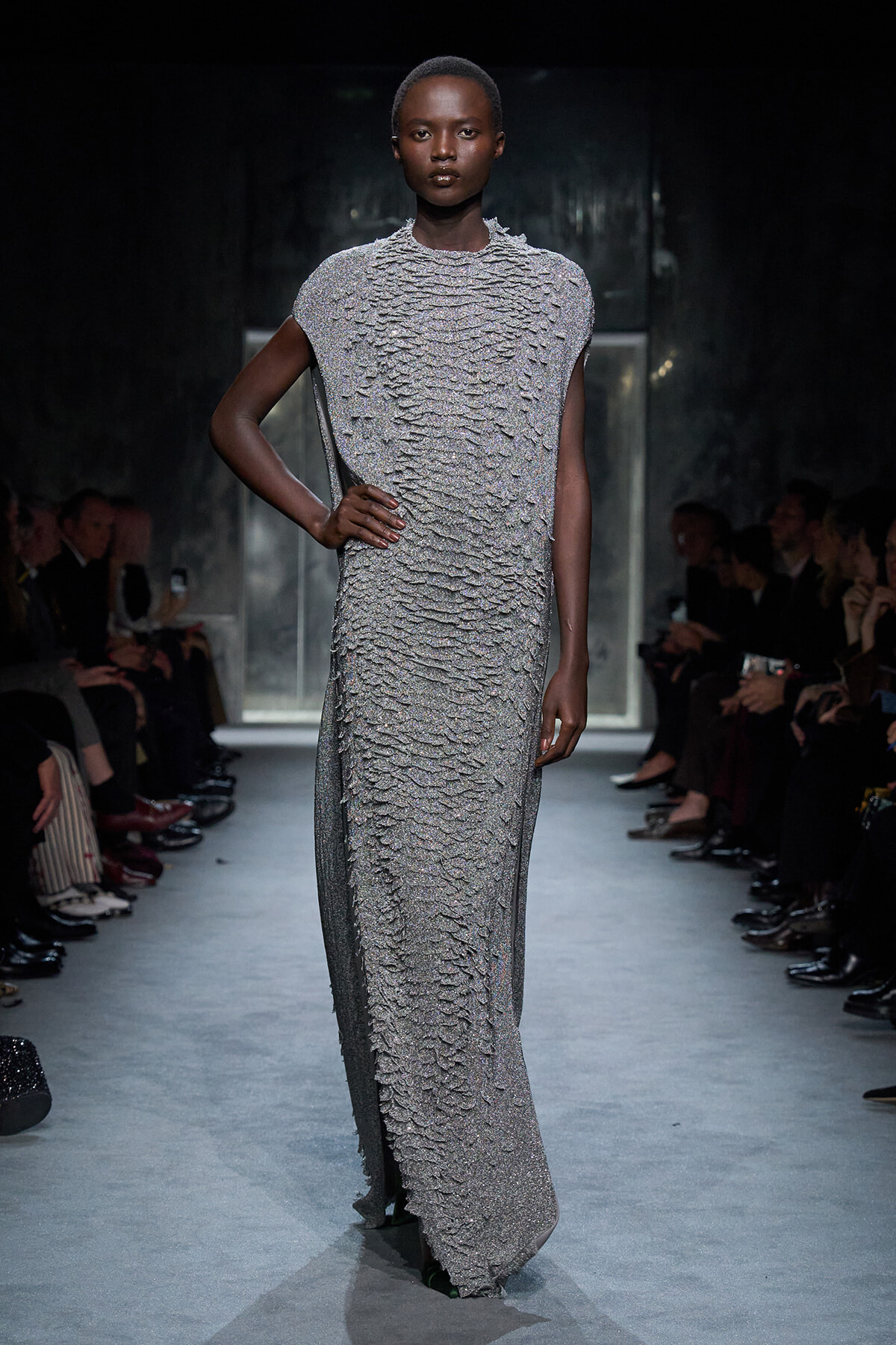 Model on a runway wearing a shimmering, textured silver floor-length gown with short sleeves, audience lining the catwalk on both sides.