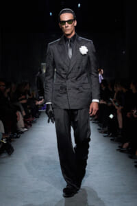 Male model walking a fashion runway in a dark dotted double-breasted suit with a white boutonniere, sunglasses, and gloves.