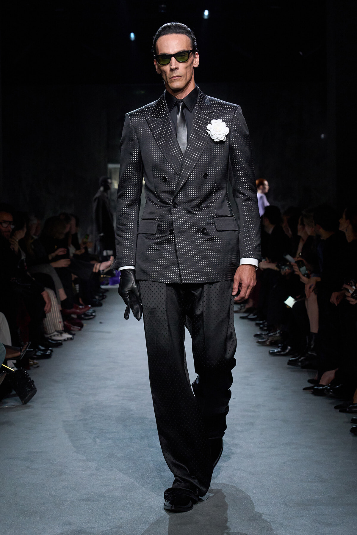 Male model walking a fashion runway in a dark dotted double-breasted suit with a white boutonniere, sunglasses, and gloves.