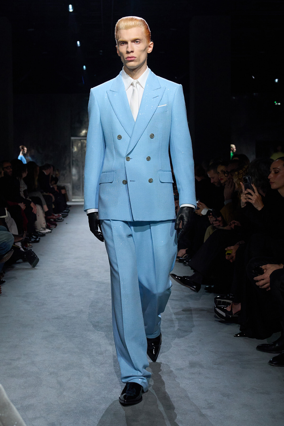 Male model walks the runway in a powder-blue double-breasted suit with a white shirt and tie, black gloves, and polished shoes. Pro backstage audience visible on sides.