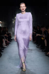Model walking a runway in a lavender fringe knit gown with long fringe sleeves and green strappy sandals, audience in background