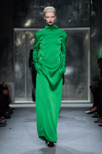 Model walking a runway in a floor-length emerald green satin gown with a high neckline and long sleeves.