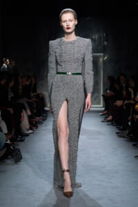 Model walks the runway in a textured silver-gray gown with a high front slit, cinched by a green belt and bold red lips.