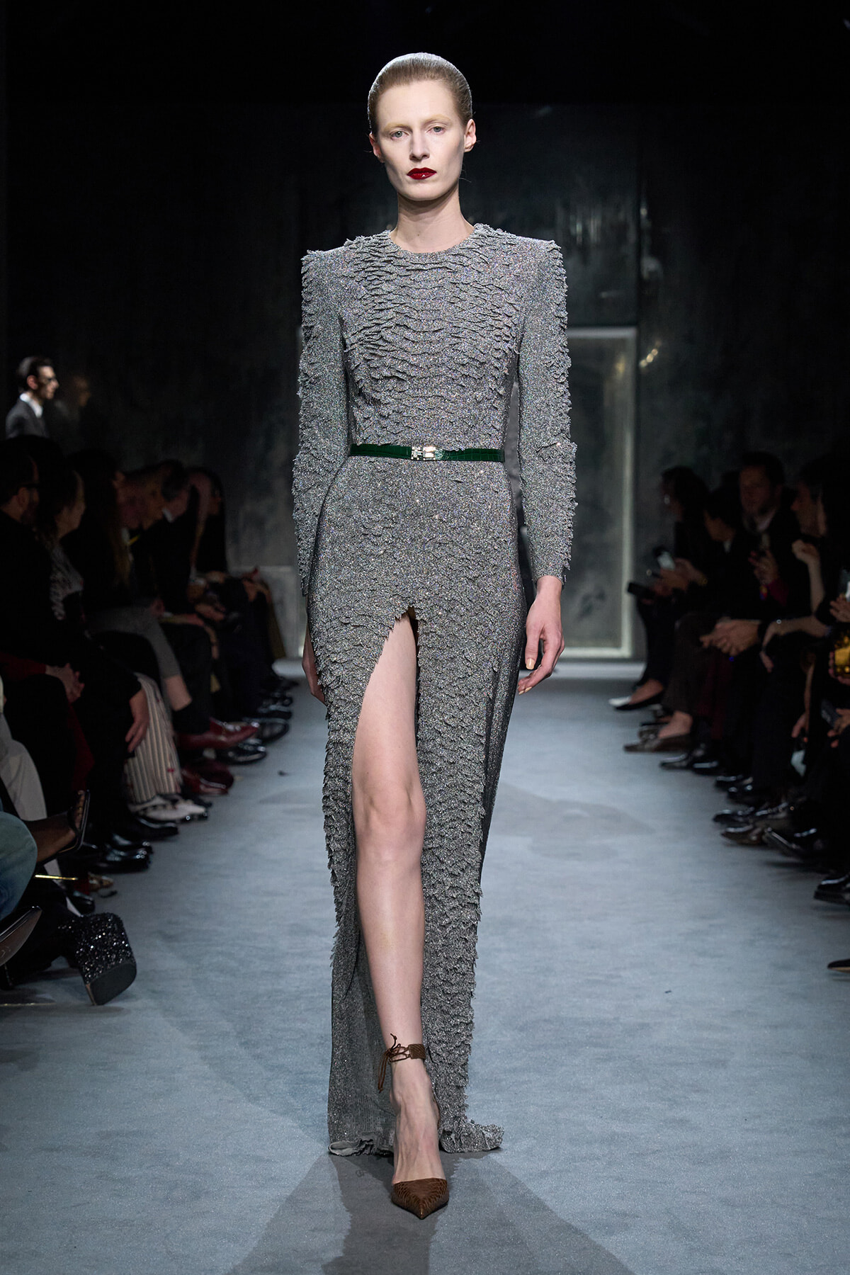 Model walks the runway in a textured silver-gray gown with a high front slit, cinched by a green belt and bold red lips.