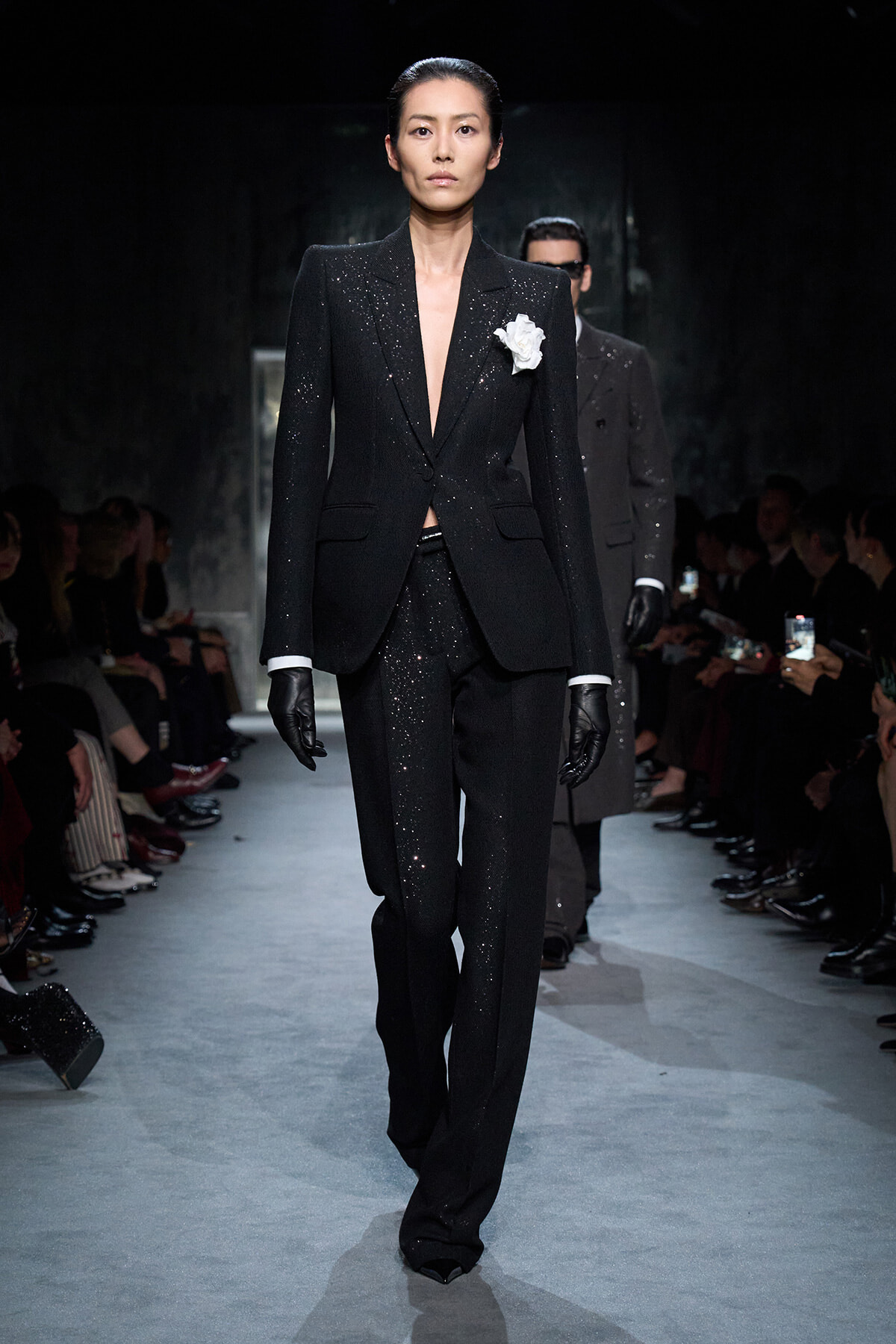 Model walking the runway in a sparkling black pantsuit with a white flower brooch and black gloves.