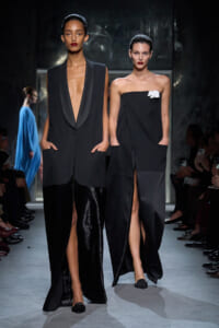 Two models walk a runway in chic black ensembles; one wears a deep V blazer with wide, tuxedo-like lapels and shimmering black pants, hands in pockets.