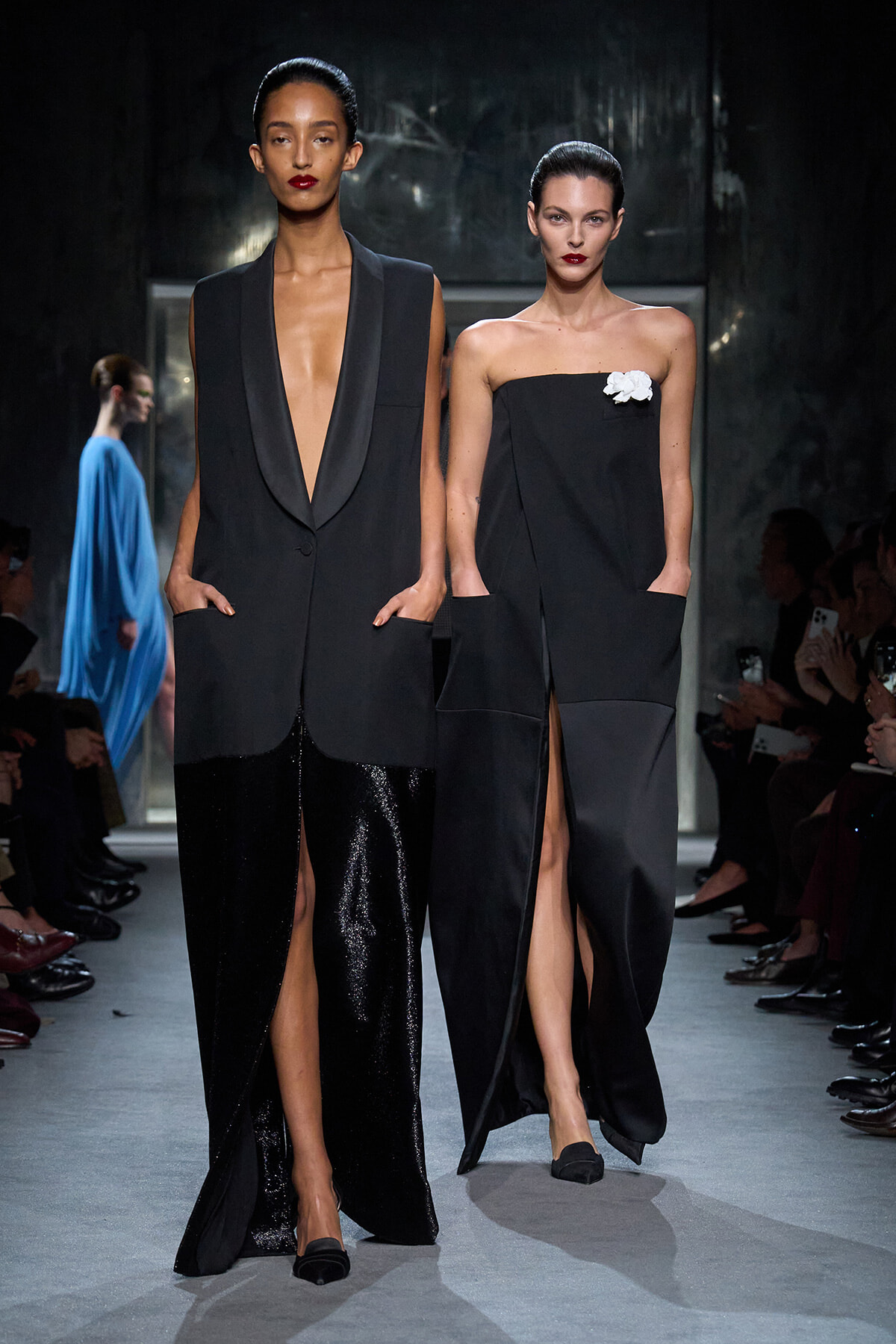 Two models walk a runway in chic black ensembles; one wears a deep V blazer with wide, tuxedo-like lapels and shimmering black pants, hands in pockets.