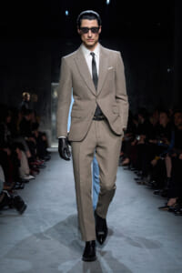 Male model in a sparkling beige suit, white shirt, black tie, and sunglasses walks the runway with black gloves.