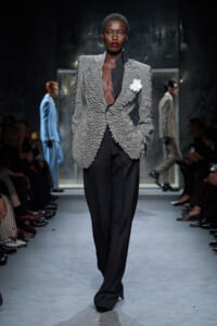 Model walks the runway in a textured gray blazer with black lapels, black wide-leg pants, and a white flower brooch.