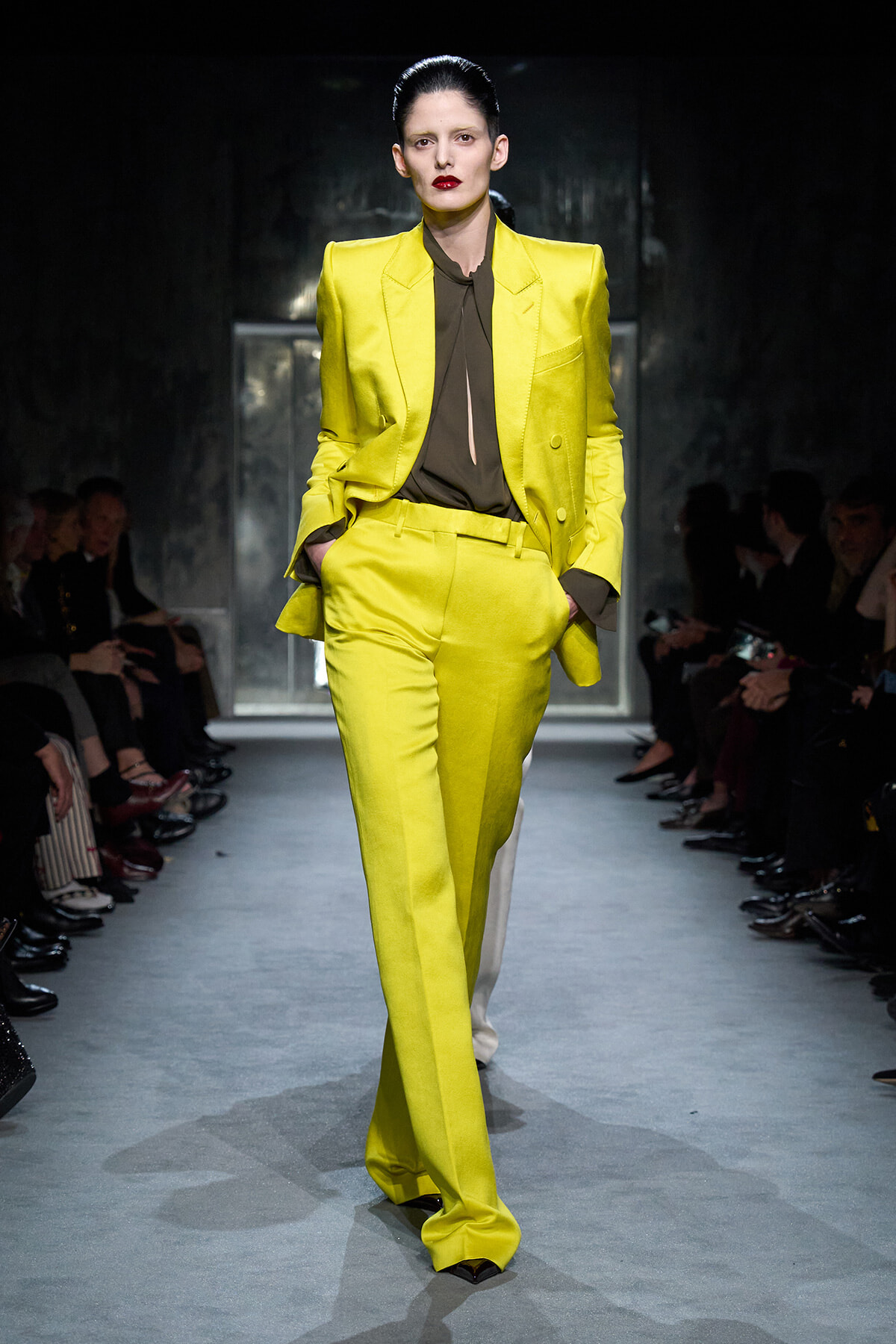 Model walking a runway in a bright yellow tailored suit with a brown blouse, hands in pockets, dark backdrop behind.