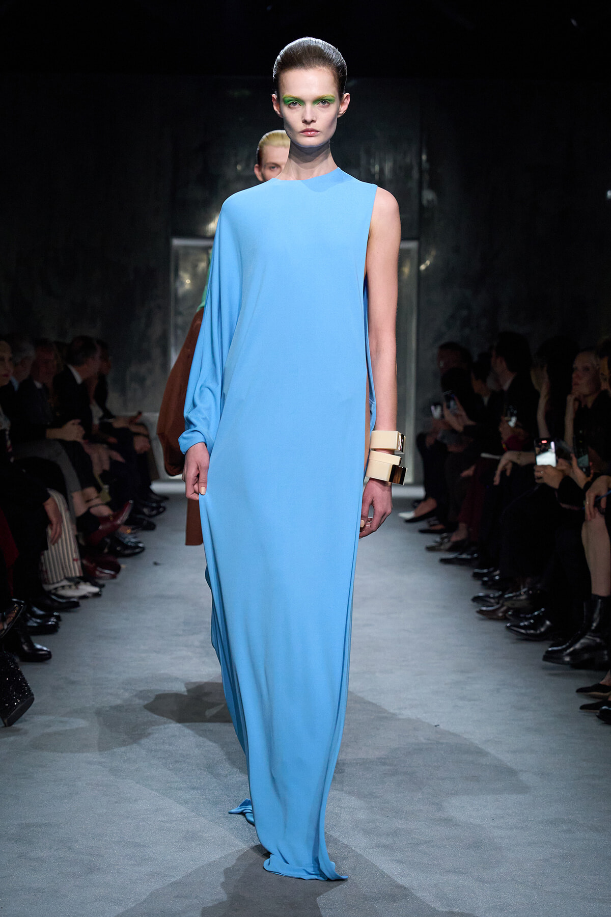 Model walks the runway in a sky-blue, one-shoulder draped gown with a long sleeve on the left and a large beige cuff bracelet, audience beside the catwalk.
