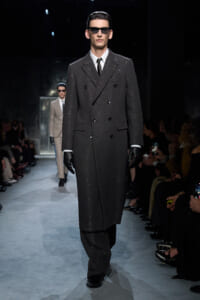 Male model walks a fashion runway in a dark, glittery double-breasted overcoat with black gloves and sunglasses.