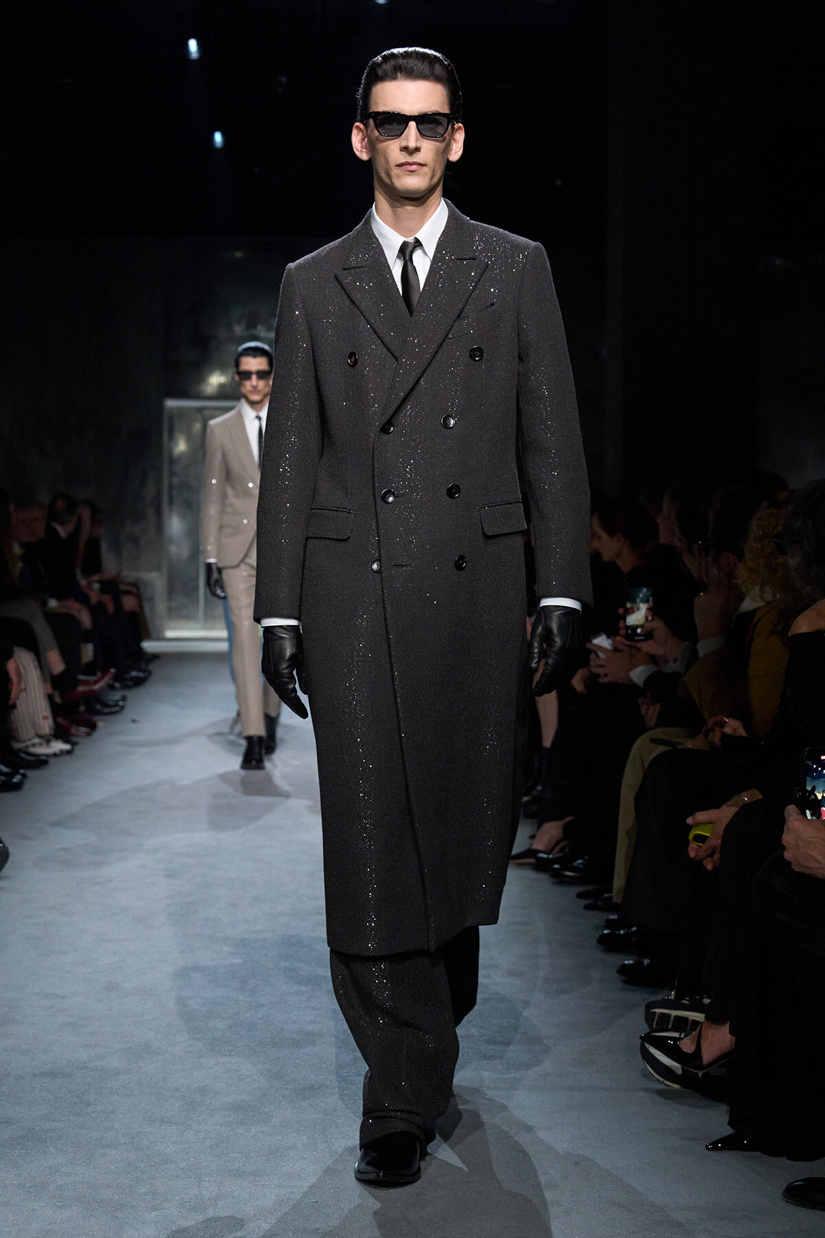 Male model walks a fashion runway in a dark, glittery double-breasted overcoat with black gloves and sunglasses.