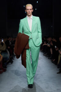 Male model walking a fashion show runway in a mint green suit, holding a brown coat over his forearm while audiences watch.