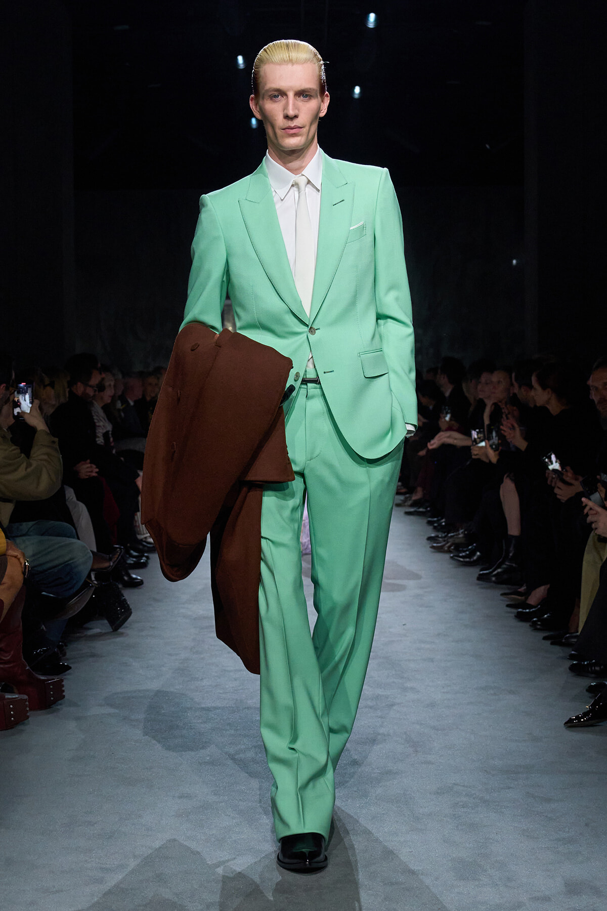 Male model walking a fashion show runway in a mint green suit, holding a brown coat over his forearm while audiences watch.