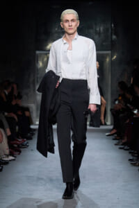 Male model walks a runway in a white dress shirt and black trousers, carrying a black coat over his arm.