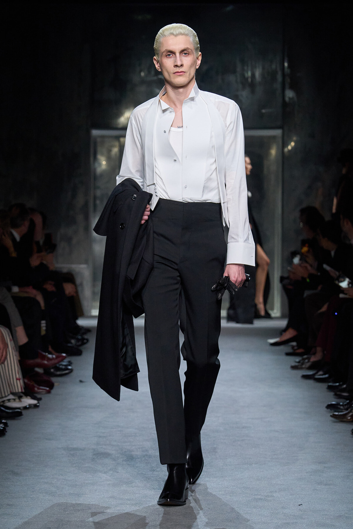Male model walks a runway in a white dress shirt and black trousers, carrying a black coat over his arm.