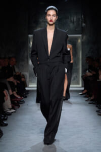 Model walks the runway wearing a tailored black suit with a deep plunging lapel, hands in pockets, red lips, slicked-back hair.