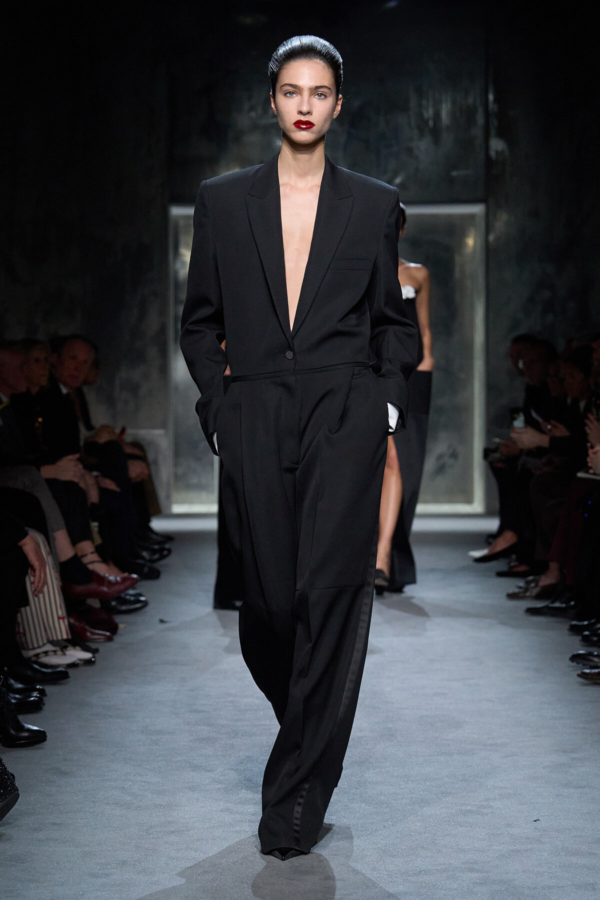 Model walks the runway wearing a tailored black suit with a deep plunging lapel, hands in pockets, red lips, slicked-back hair.