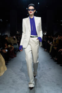 Model on a runway wearing a white tailored suit with a cobalt blue blouse and dark sunglasses, bold red lips.