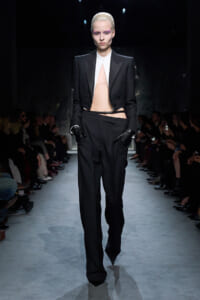 Fashion runway image: a model wears a black tailored pantsuit with a cropped blazer, white shirt, peach centerpiece, and black gloves. Audience on sides.