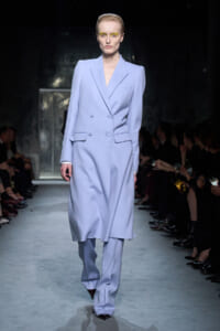 Model on a fashion runway wearing a pale lavender oversized double-breasted blazer with matching wide-leg pants and bold yellow eye makeup.