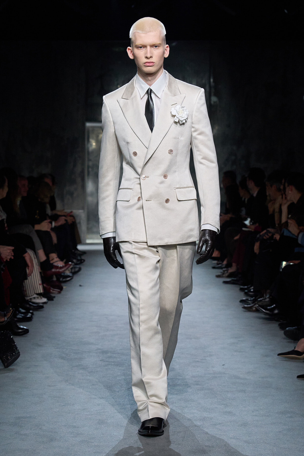 Male model walking a white double-breasted suit with a black tie and lapel flower, on a fashion runway.