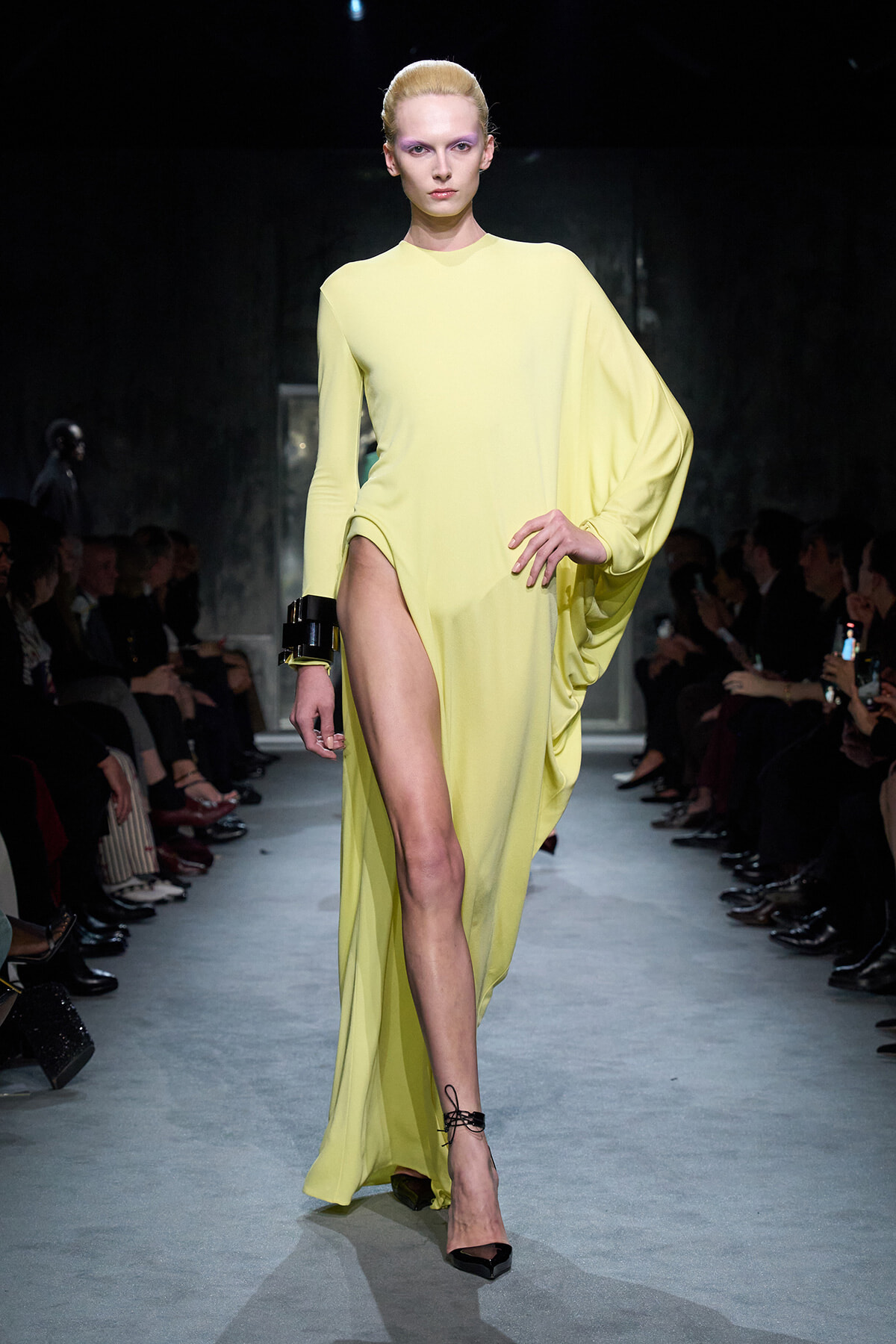 Model walks the runway in a pale yellow, one-shoulder, floor-length gown with a high thigh slit and black bracelet accessory on the left wrist