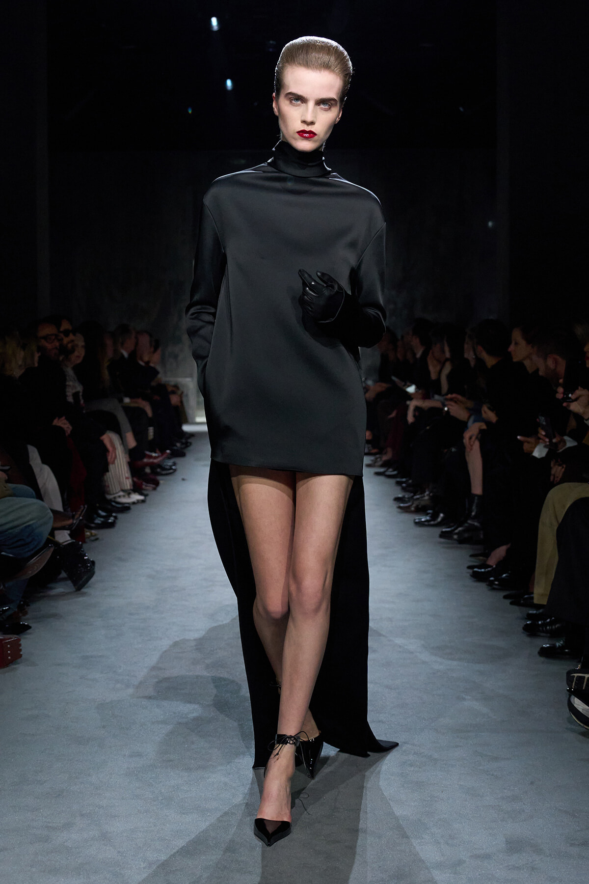 Model walks a dark runway in a black high-neck mini dress with a cape-like back, black gloves, and pointed heels.