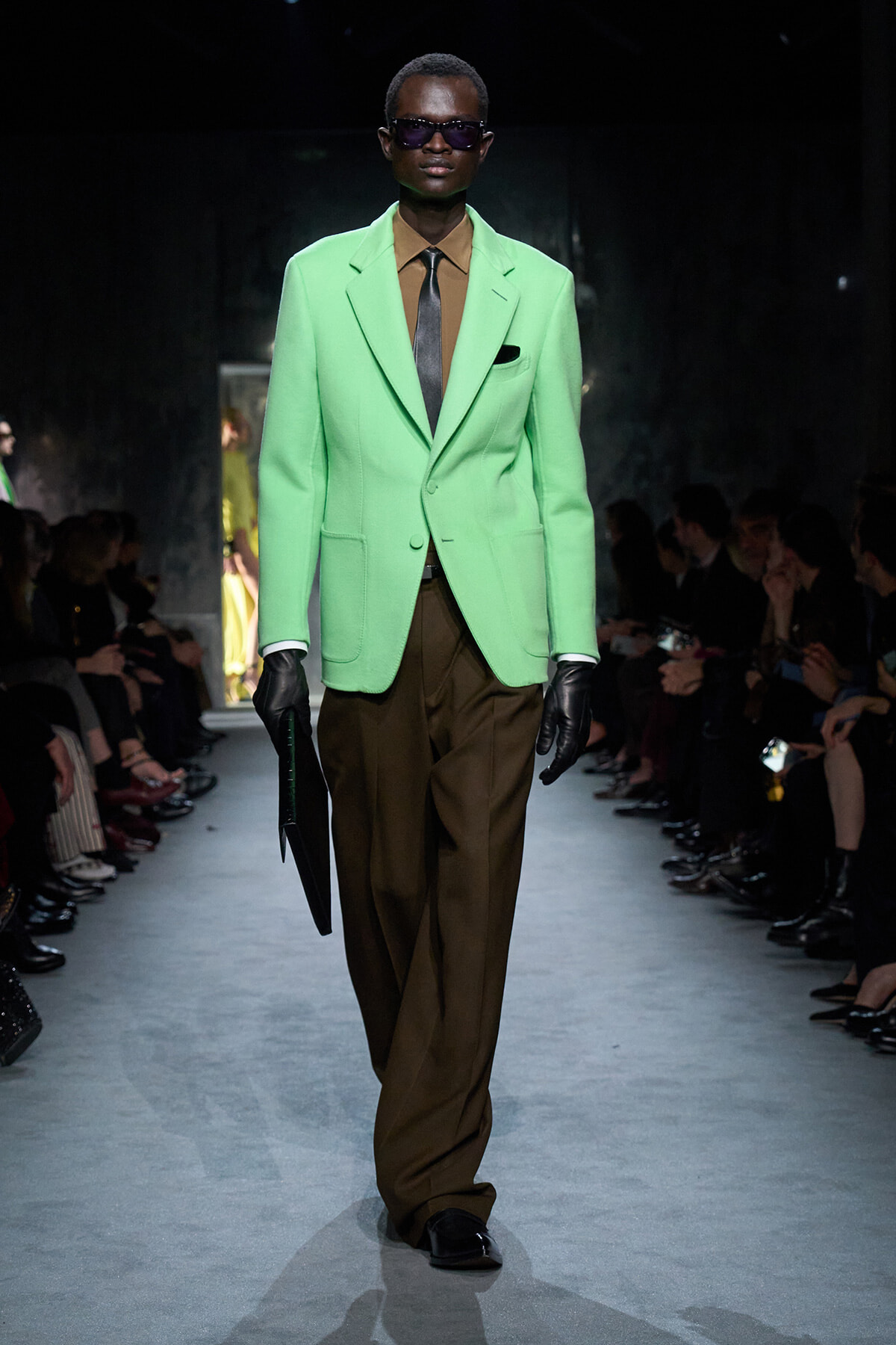 Model walking a runway in a lime-green blazer, brown shirt and tie, with dark sunglasses and black gloves while carrying a slim black folder.