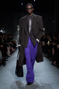 Male model on a runway wearing a dark brown oversized blazer, charcoal suit, silver tie, and vibrant purple trousers while carrying a black textured bag and wearing sunglasses.