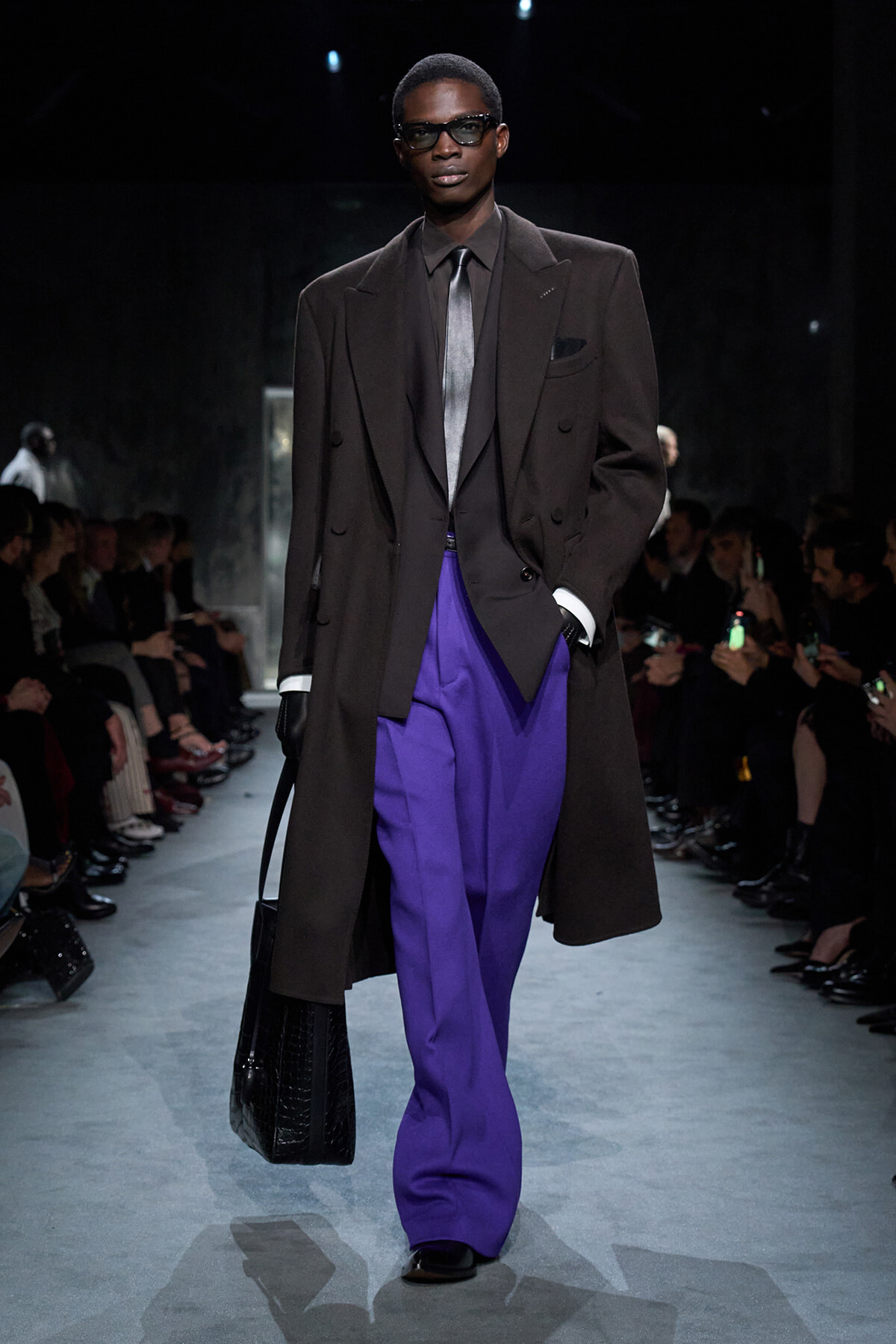 Male model on a runway wearing a dark brown oversized blazer, charcoal suit, silver tie, and vibrant purple trousers while carrying a black textured bag and wearing sunglasses.