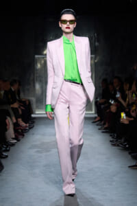 Model on a runway wearing a pastel pink suit with a neon green blouse and dark sunglasses, red lipstick.